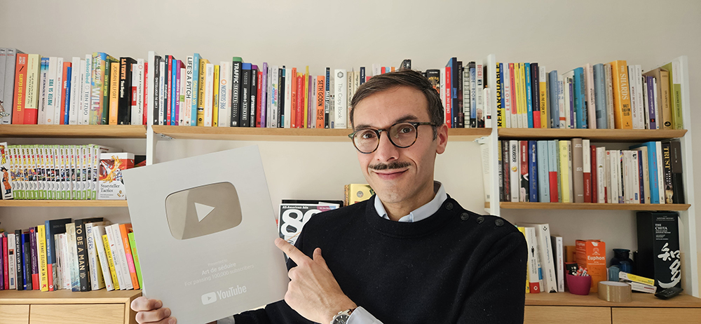 Sélim Niederhoffer montrant sa récompence Youtube, porte un pull noir et livres en fond